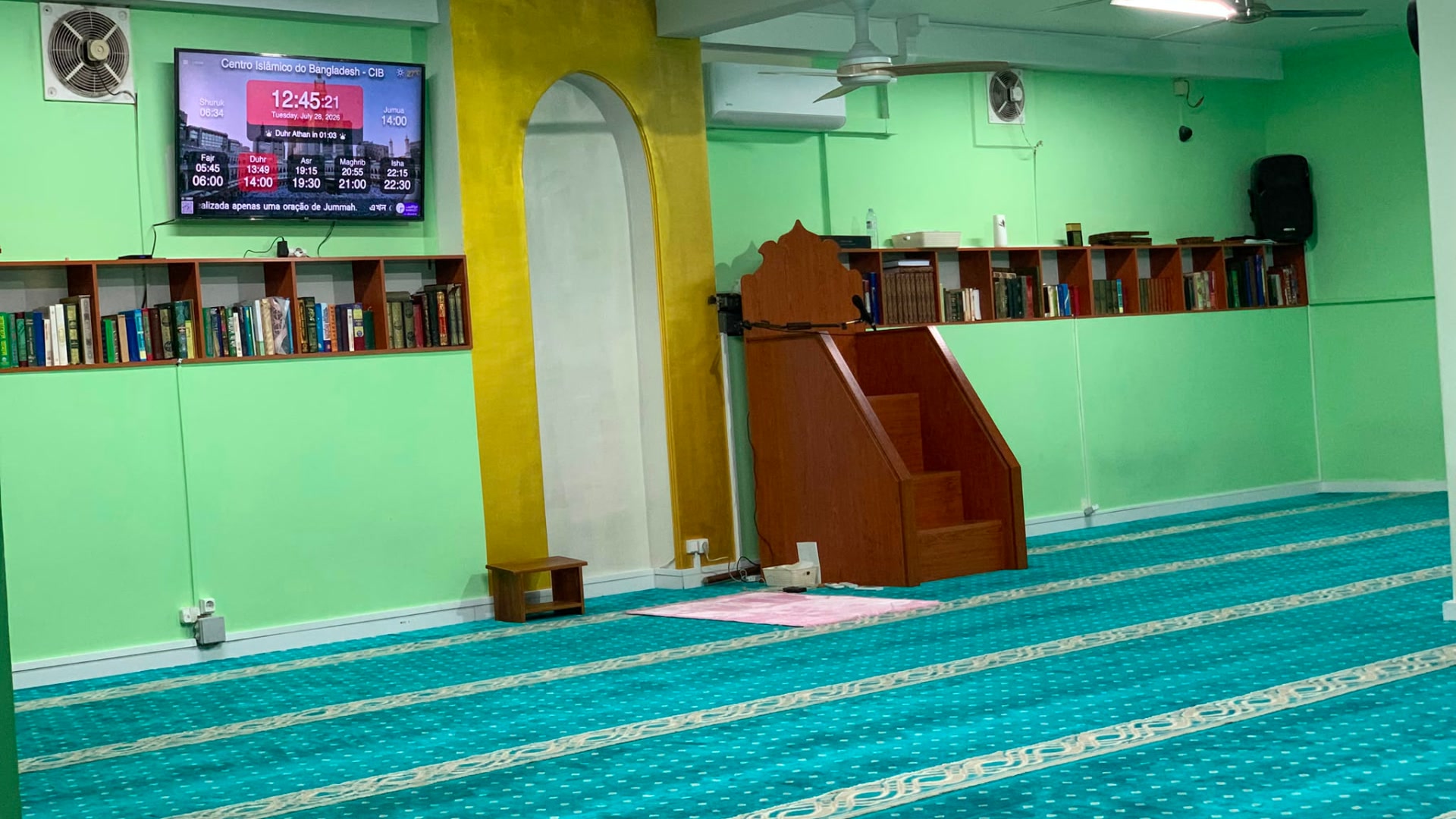 Interior of the prayer hall with teal carpets, bookshelves, and a wooden minbar
